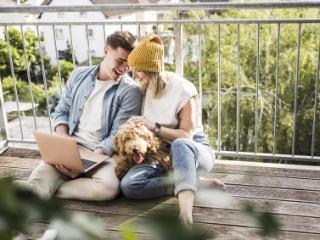 Junges Paar sitzt mit dem Hund auf dem Balkon und schaut in den Laptop